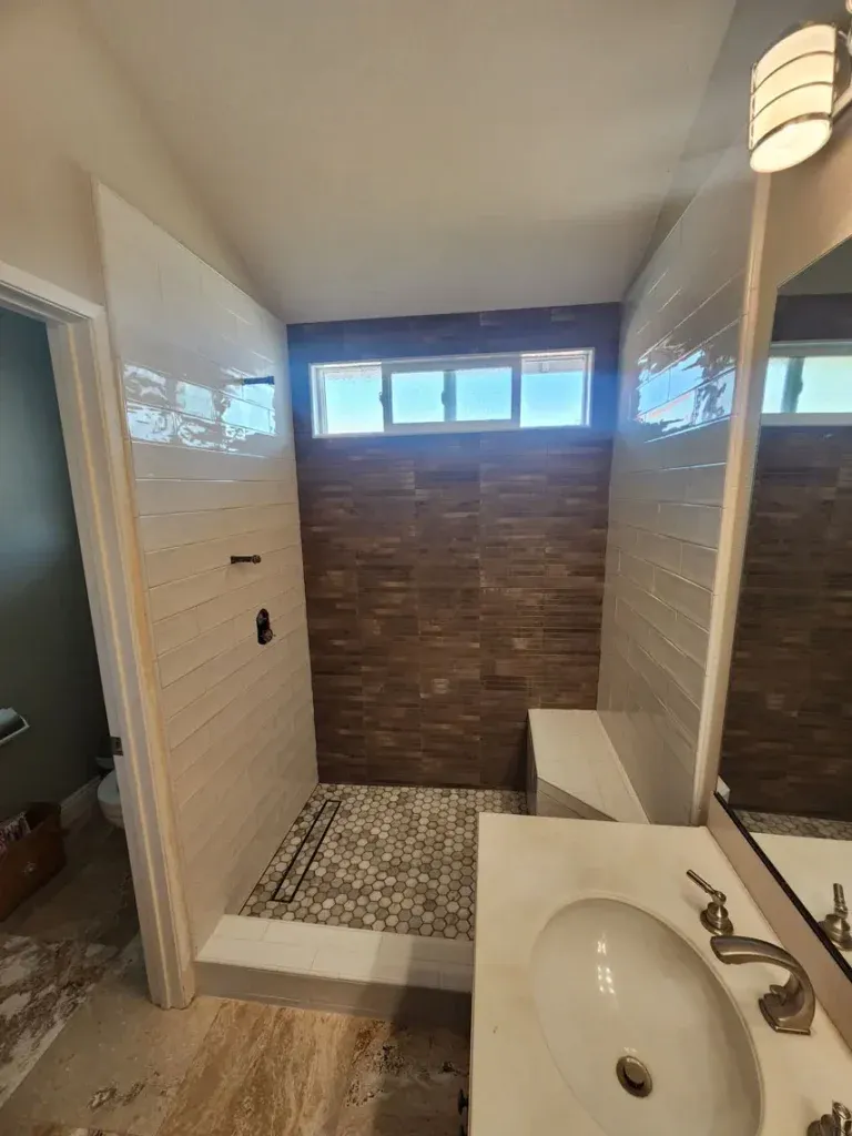 Fort Mohave bathroom remodel with walk-in shower, brown and white tiles, window, white sink, wall light, and vinyl plank flooring.