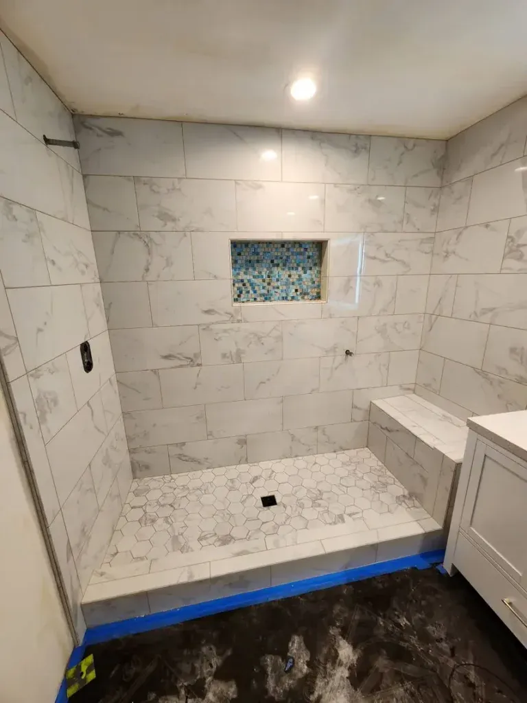 Shower under construction in Bullhead City with white marble tiles, hex floor, bench, blue niche, and expert tile work.